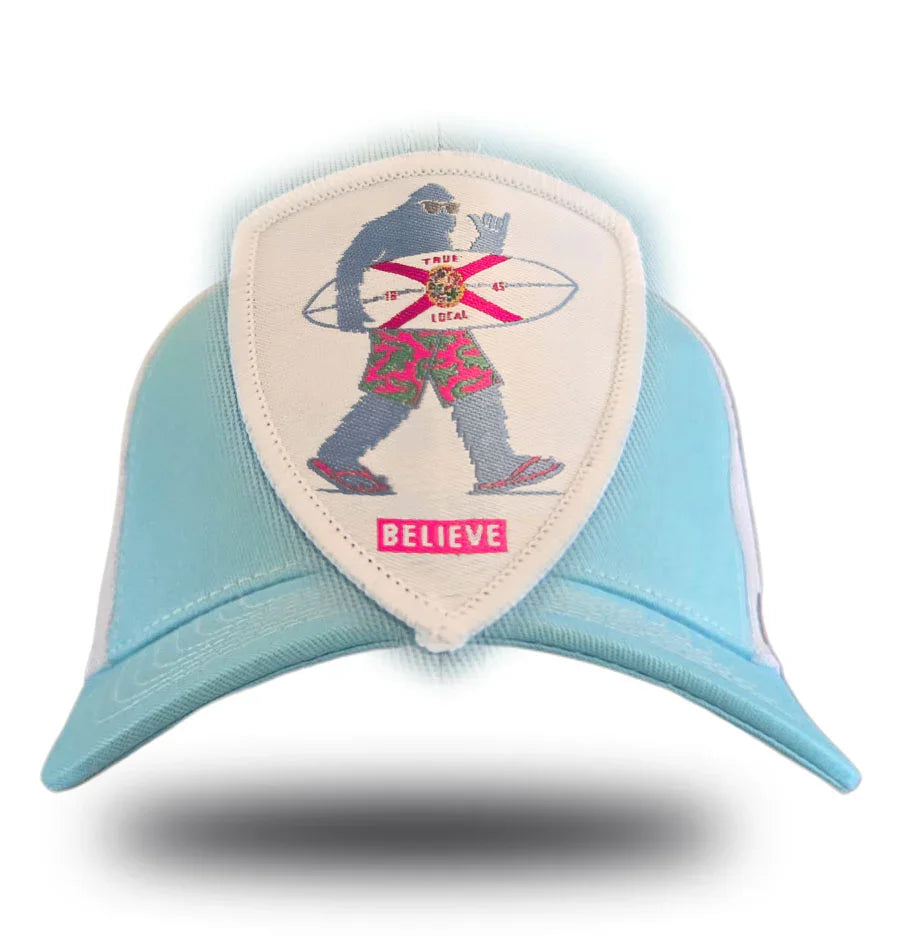 Light blue trucker hat with embroidered Bigfoot holding surfboard, Florida flag, 'BELIEVE' patch, local pride apparel