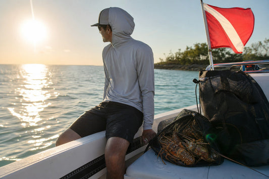 Diver in a gray dri-fit hooded long sleeve sitting on a boat gunwale at sunset with a mesh bag of spiny lobster and a red dive flag, Florida Keys
