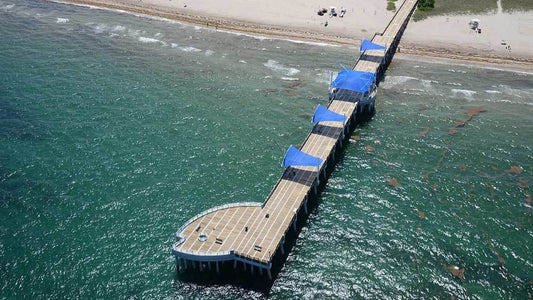 Pompano Beach Fisher Family Pier — aerial view of the renovated 900-foot pier with Pompano fish-shaped end and blue sail shades over turquoise Atlantic water